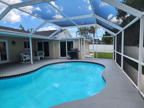 A swimming pool with a curved edge, surrounded by a covered patio area with chairs and a grill.