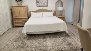 Un dormitorio con una cama grande, un armario de madera, una mesita de noche con una lámpara, un ventilador de techo, un cuadro y una ventana con cortinas.