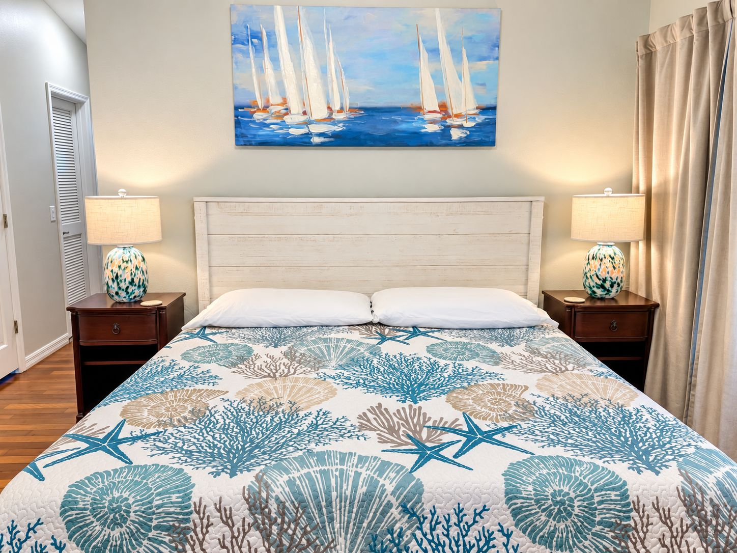 Habitación de hotel con cama, dos mesitas de noche, un cuadro de veleros y vista al mar.