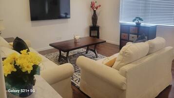 A living room with a flat-screen TV, a coffee table, two sofas, a vase with flowers, a clock, and a window with blinds.