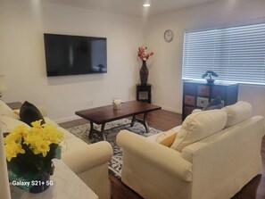 A living room with a flat-screen TV, a coffee table, two sofas, a vase with flowers, a clock, and a window with blinds.