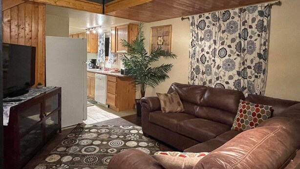 A cozy living room with a brown leather sofa, a television, and a patterned rug. A kitchen is visible in the background with wooden cabinets and a potted plant. - Sled Dog Cabin: Family, Pet Friendly Quiet Home in Wasilla (Wasilla)