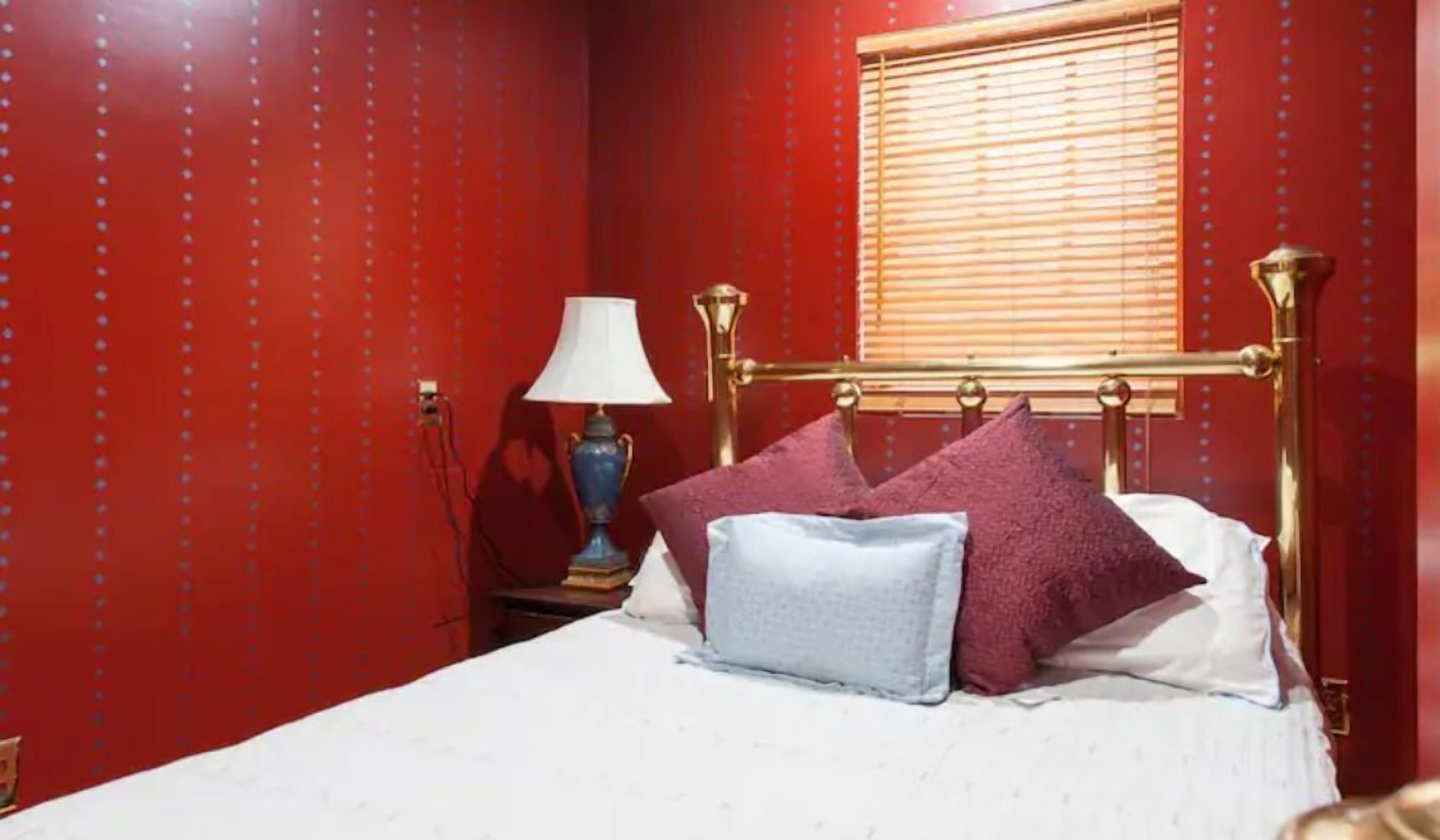 A room with a bed, red walls, a window with blinds, a nightstand with a lamp, and a blue lamp.