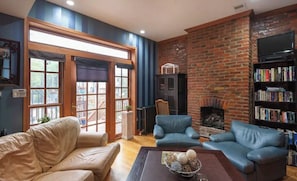 A cozy living room with a brick fireplace, leather sofas, a bookshelf, and a large window.