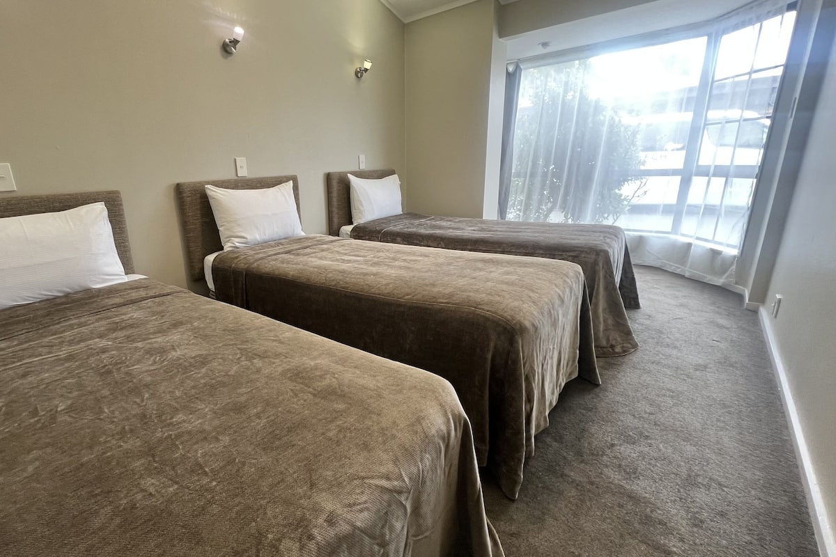 A hotel room with three beds, each with a brown bedspread and white pillows. The room has a large window with sheer curtains, allowing natural light to enter.