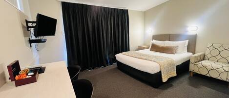 A hotel room with a bed, bedside table, chair, and a desk with a computer and some items on it.
