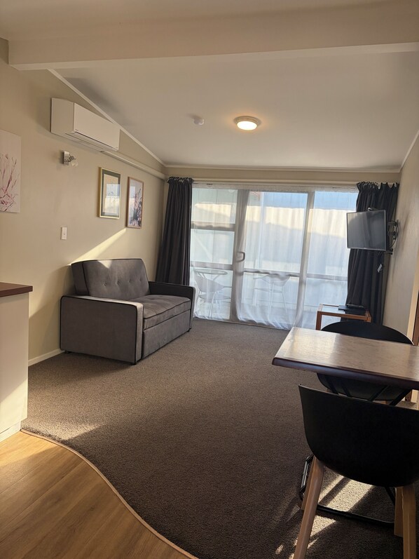 A living room with a grey sofa, a small table, a TV, and a sliding glass door leading to a balcony. - Marineland Motel (Napier)
