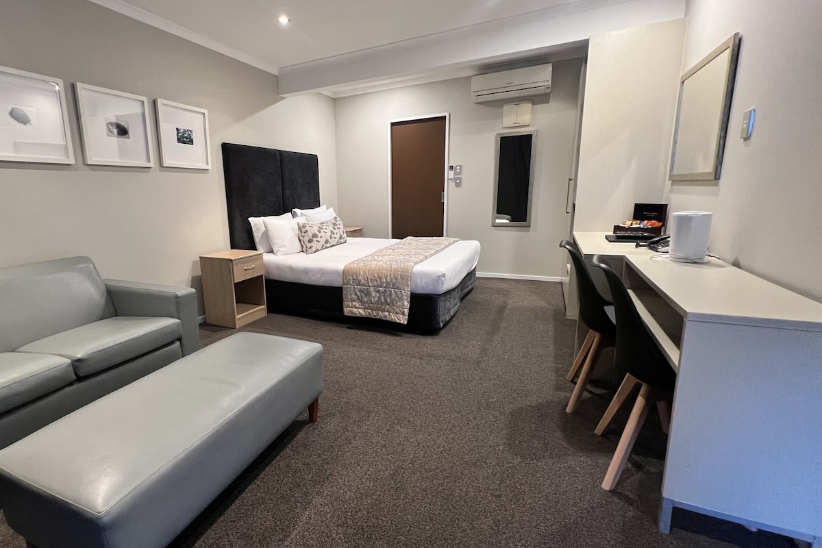 A modern hotel room with a grey sofa, a bed with a white duvet, a desk with a chair, and a small bedside table.