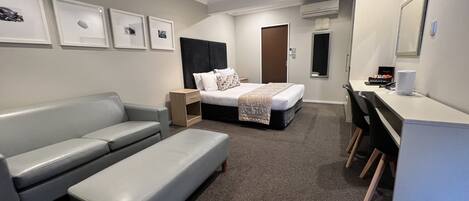 Et moderne hotelværelse med en grå sofa, en seng med hvid dyne, et skrivebord med stol, og et lille natbord.