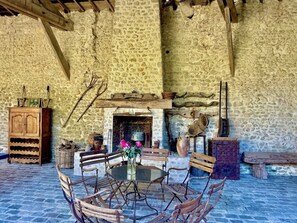 A rustic stone interior with a round table, wooden chairs, a fireplace, and a wooden cabinet.