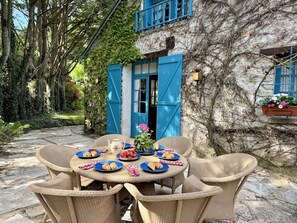 Une table ronde entourée de chaises en osier, un chemin de pierres et un bâtiment aux volets bleus.