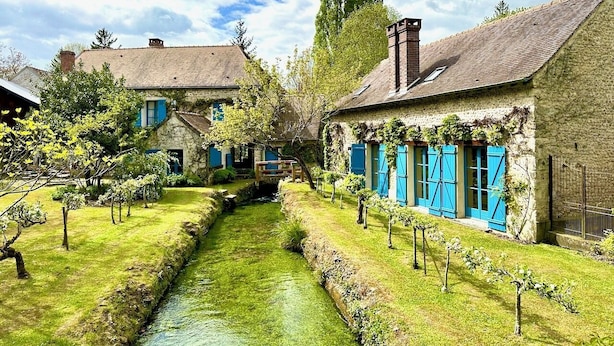 A narrow water channel between buildings with a rustic architectural style, a small bridge, and a well-maintained grassy area. - Elegant Mill Compound, 1 hr from Paris. Sleep 20, Events for 50 - an extra fee. (Méréville)