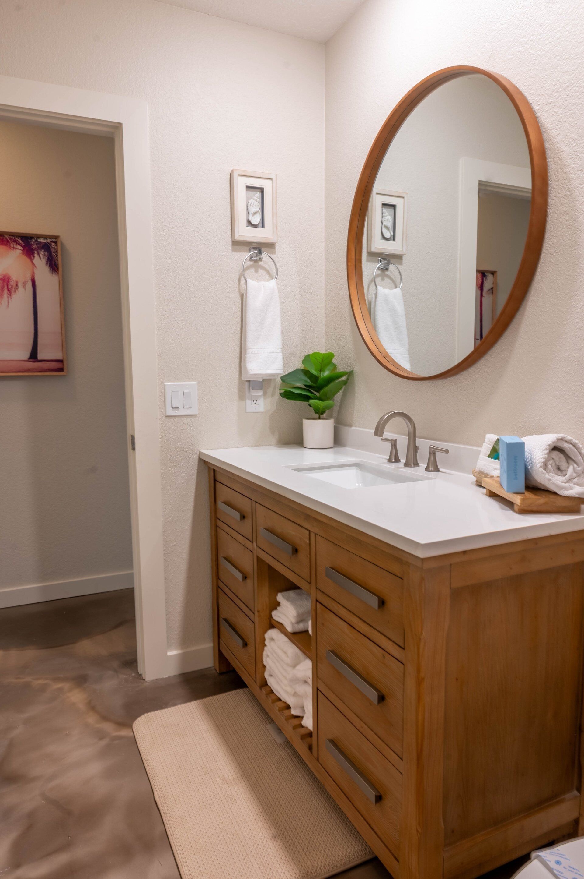 A bathroom with a wooden vanity, a white sink, a mirror, a potted plant, and a painting on the wall.