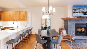 A modern kitchen with wooden cabinets, a central island with bar stools, a chandelier, and a fireplace with a TV above it. - 2BR ski-in/out condo in Beaver Creek Village with mountain views (Beaver Creek)