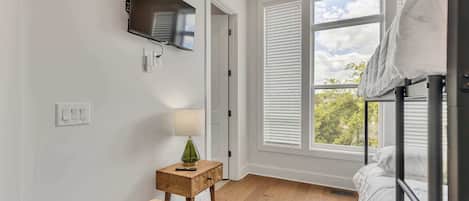 A bedroom with a bed, a TV mounted on the wall, a small table with a lamp, and a window with blinds.