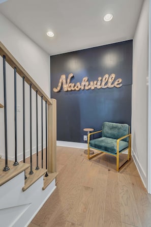 A staircase with wooden steps and black balusters, a dark wall with a neon sign reading "Nashville," and a green upholstered chair with gold legs.