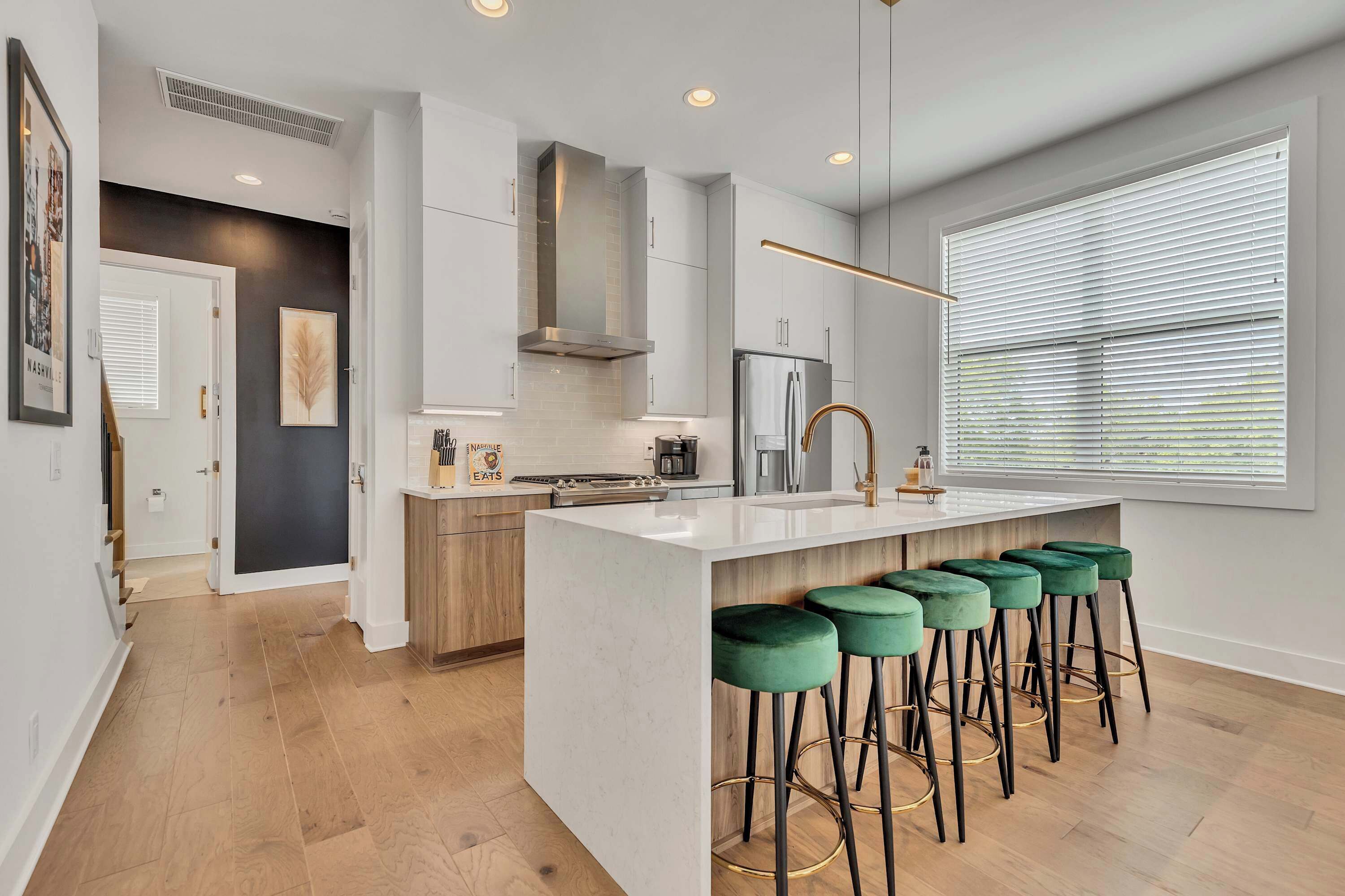 A modern kitchen with a central island, bar stools, and a large window with blinds.