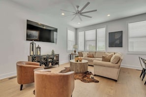A modern living room with a ceiling fan, a large flat-screen TV, a beige sofa, two leather armchairs, a round wooden coffee table, and a cowhide rug. - New Nashville Gem | Rooftop & Sleeps 11 (Nashville)