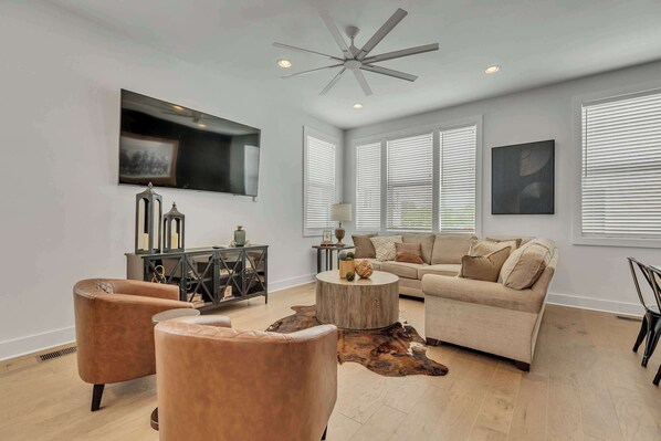 A modern living room with a ceiling fan, a large flat-screen TV, a beige sofa, two leather armchairs, a round wooden coffee table, and a cowhide rug.