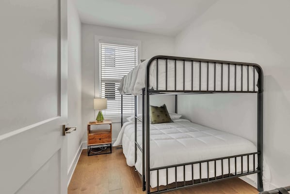 A small, single-bed room with a bunk bed, a wooden nightstand with a lamp, and a window with blinds.