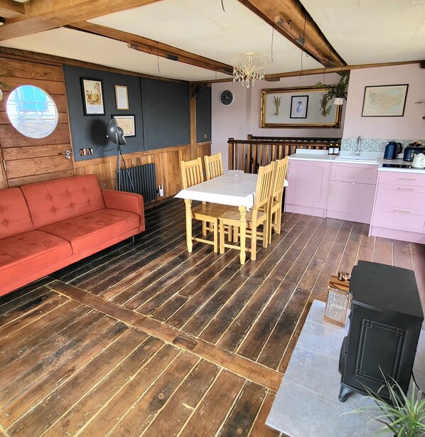 A cozy room with a wooden floor, a red sofa, a dining table with chairs, a kitchen area with pink cabinets, and a chandelier.
