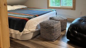A bedroom with a sloped ceiling, a large window, a bed with a colorful blanket, two woven ottomans, and a black leather bench. - Trail House, Denali Brewing, Netflix, Concert, Fire Pit, Family, Race, festival  (Talkeetna)