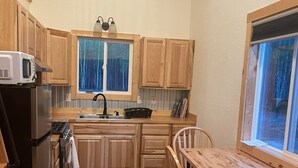 A compact kitchen with wooden cabinets, a built-in microwave, a sink, and a dining table with chairs. - Trail House, Denali Brewing, Netflix, Concert, Fire Pit, Family, Race, festival  (Talkeetna)