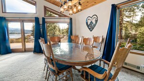 Un coin repas avec une table ronde en verre entourée de chaises en bois dotées de coussins bleus, de grandes fenêtres avec des rideaux bleus, et une vue sur un lac et des montagnes.