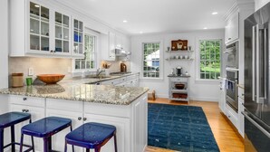 Una cocina amplia con armarios blancos, encimeras de granito, una isla grande con taburetes y vistas al exterior a través de una ventana.