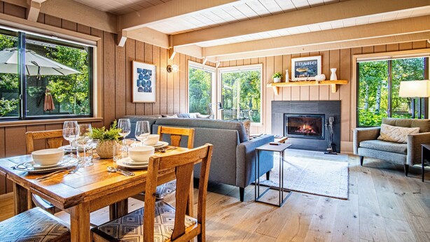 A cozy living room with a fireplace, wooden floors, and large windows offering a view of greenery. - Wildwood Cottage at Villager + SV Resort Amenities (Sun Valley)