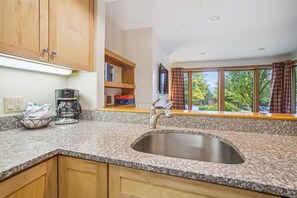 A kitchen with granite countertops, a stainless steel sink, wooden cabinets, a coffee maker, and a window with curtains. - 2 bedroom Ski-in condo with pool, hot tub, and EV chargers (Killington)