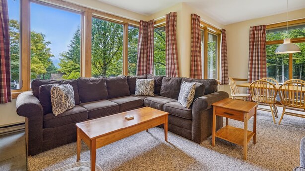 A cozy living room with a sectional sofa, a wooden coffee table, and a dining table with chairs. Large windows offer a view of trees and a clear sky. - 2 bedroom Ski-in condo with pool, hot tub, and EV chargers (Killington)