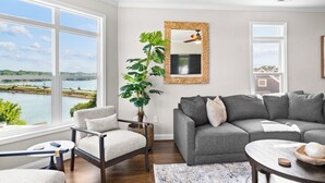 A living room with a view of a lake, a grey sofa, a chair, a table, and a large mirror on the wall. - Point of View by SoCharm | Luxury Lakefront Condo | Deck | Sleep 6 (Mooresville)