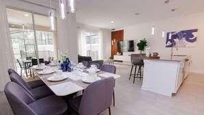 Een moderne eethoek met een grote tafel voor zes personen, omringd door stoelen. In de achtergrond is een keuken met een wit aanrecht en een televisie te zien.