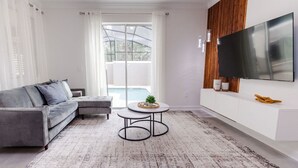 Een moderne woonkamer met een grijze bank, een kleine ronde tafel en een grote tv aan een witte muur.