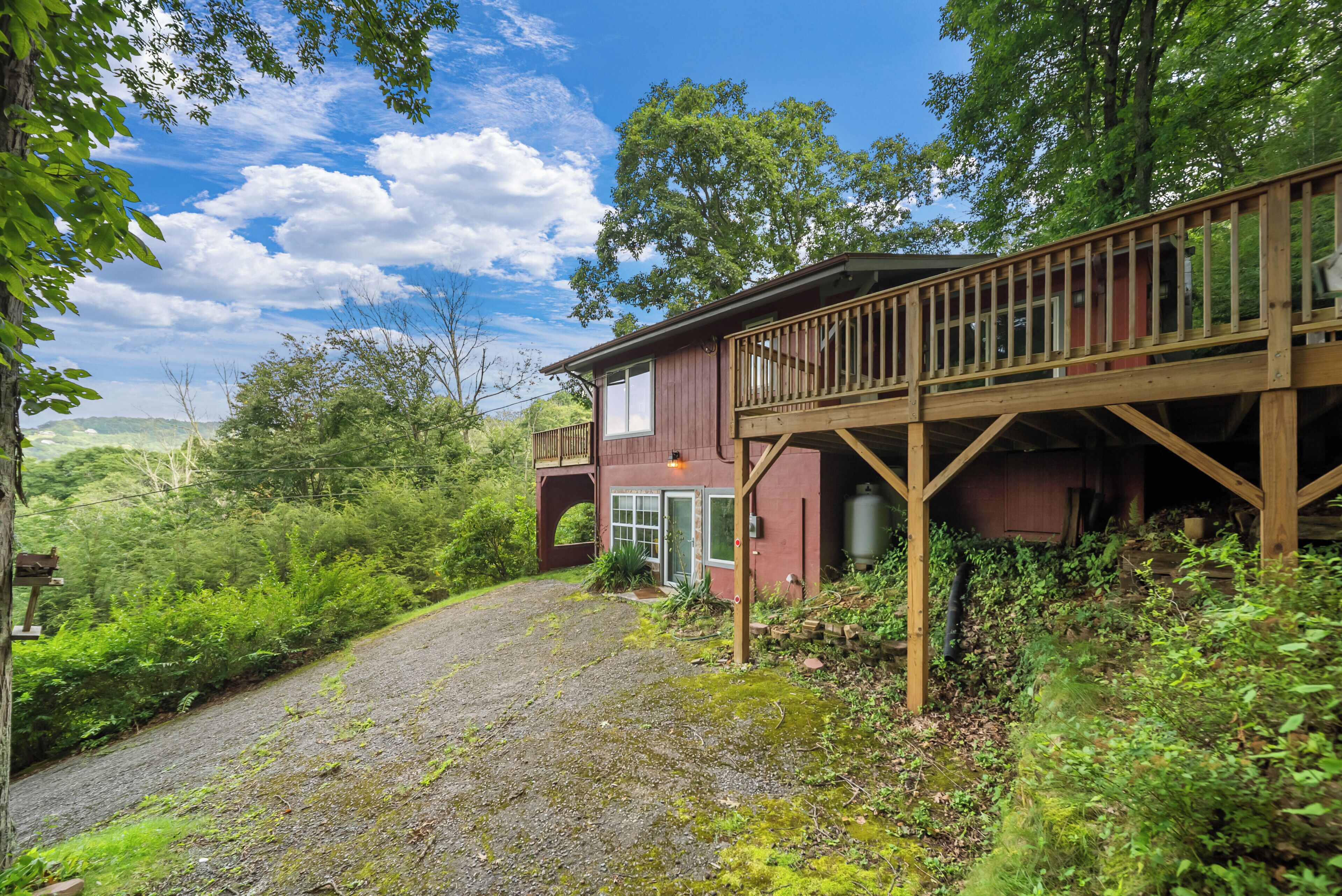 Ein zweistöckiges Holzhaus mit einer Terrasse, umgeben von üppiger Vegetation und einer Schotterauffahrt.