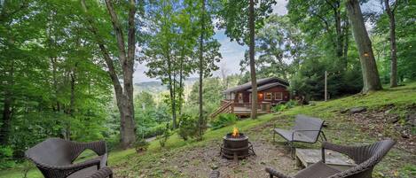 A rustic outdoor seating area with wicker chairs and a fire pit, surrounded by lush greenery and a cabin in the background.