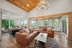 A spacious living room with a large L-shaped brown leather sofa, a white coffee table, and a wooden ceiling with a modern chandelier. - A Red Chalet - Linville mountain chalet with game room, fireplace & year-round views (Newland)