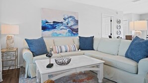 A modern living room with a light-colored sectional sofa, a coffee table, and a large abstract painting on the wall. - Sea 'n Stars Cape San Blas Vacation Rental (Port St. Joe)