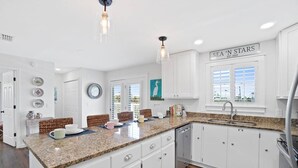 A modern kitchen with white cabinets, granite countertops, and a built-in dishwasher. There is a window with a view of the city, and a sign that reads "SEA 'N STARS EST. 2020". - Sea 'n Stars Cape San Blas Vacation Rental (Port St. Joe)