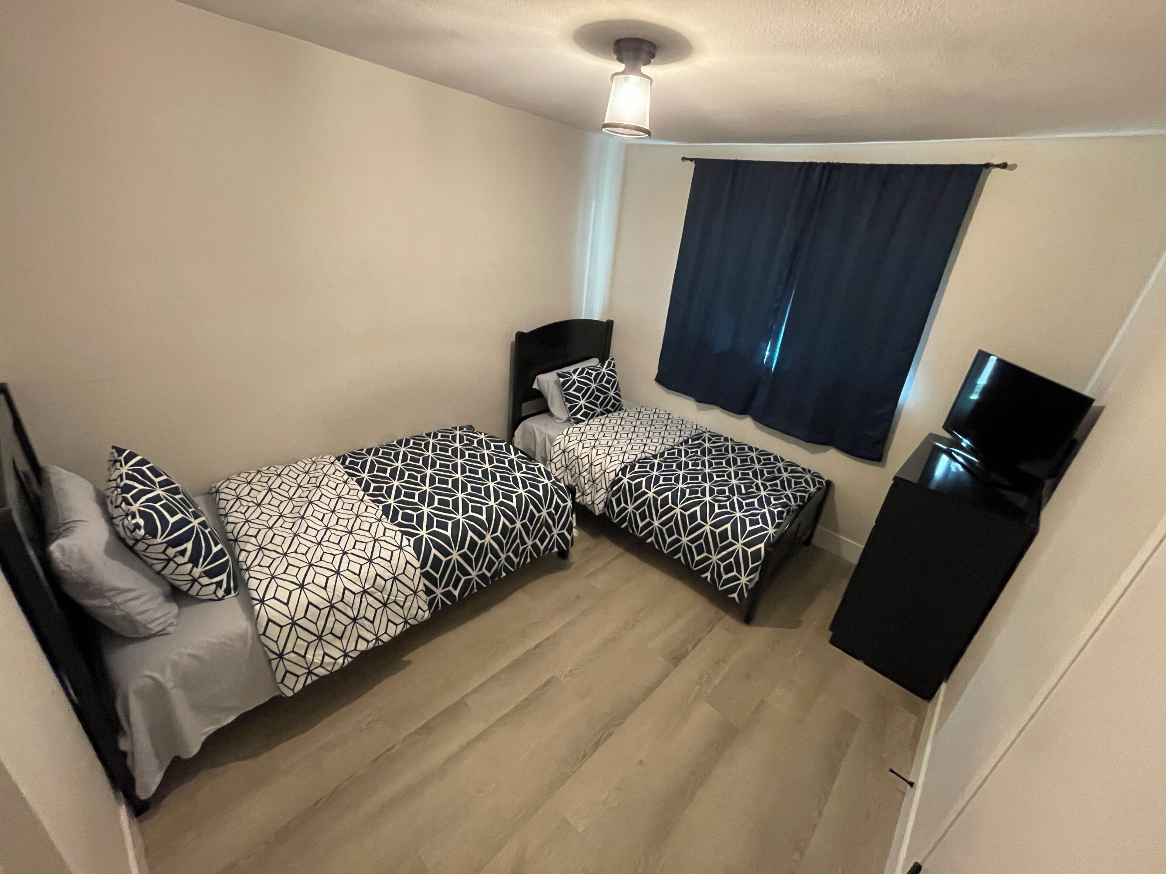 Una habitación con dos camas, un ventilador de techo, una ventana con cortinas y un mueble para televisor.