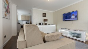 A modern living room with a beige sofa, a flat-screen TV mounted on the wall, a white entertainment unit, and a kitchen in the background.