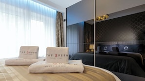 A hotel room with a bed, two towels with "The Queen Luxury Living" branding, a large window with curtains, and a modern wall design.