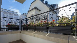 A balcony with black metal railings, potted plants, and a view of a white building with a blue roof.