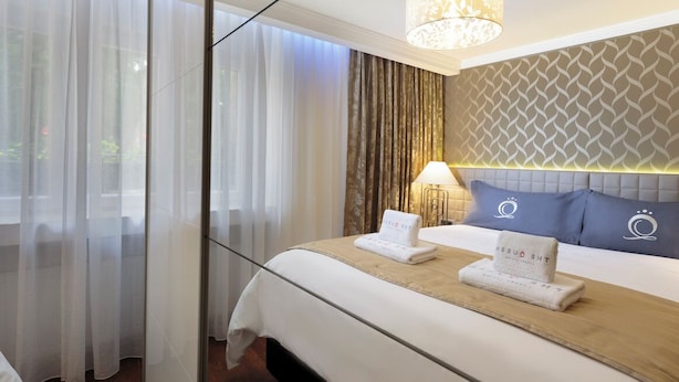 A hotel room with a large bed, a bedside table, a lamp, and a view of the outdoors through sheer curtains.
