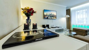 Una habitación de hotel moderna con una televisión de pantalla plana, un jarrón de flores, un cuadro enmarcado con una vista urbana, una mesa con una revista y una lámpara.