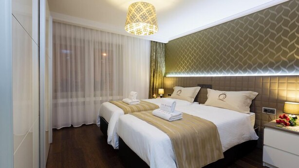 Habitación de hotel con dos camas, un ventanal con cortinas translúcidas, un papel tapiz estampado y una mesita de noche con una lámpara y flores.