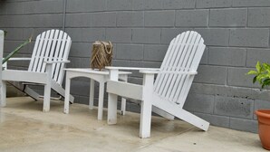 Kaksi valkoista Adirondack-tyylistä puutarhatuolia, niiden vieressä punottu kori ja terrakottasavesta valmistettu ruukku, taustalla harmaa tiiliseinä.