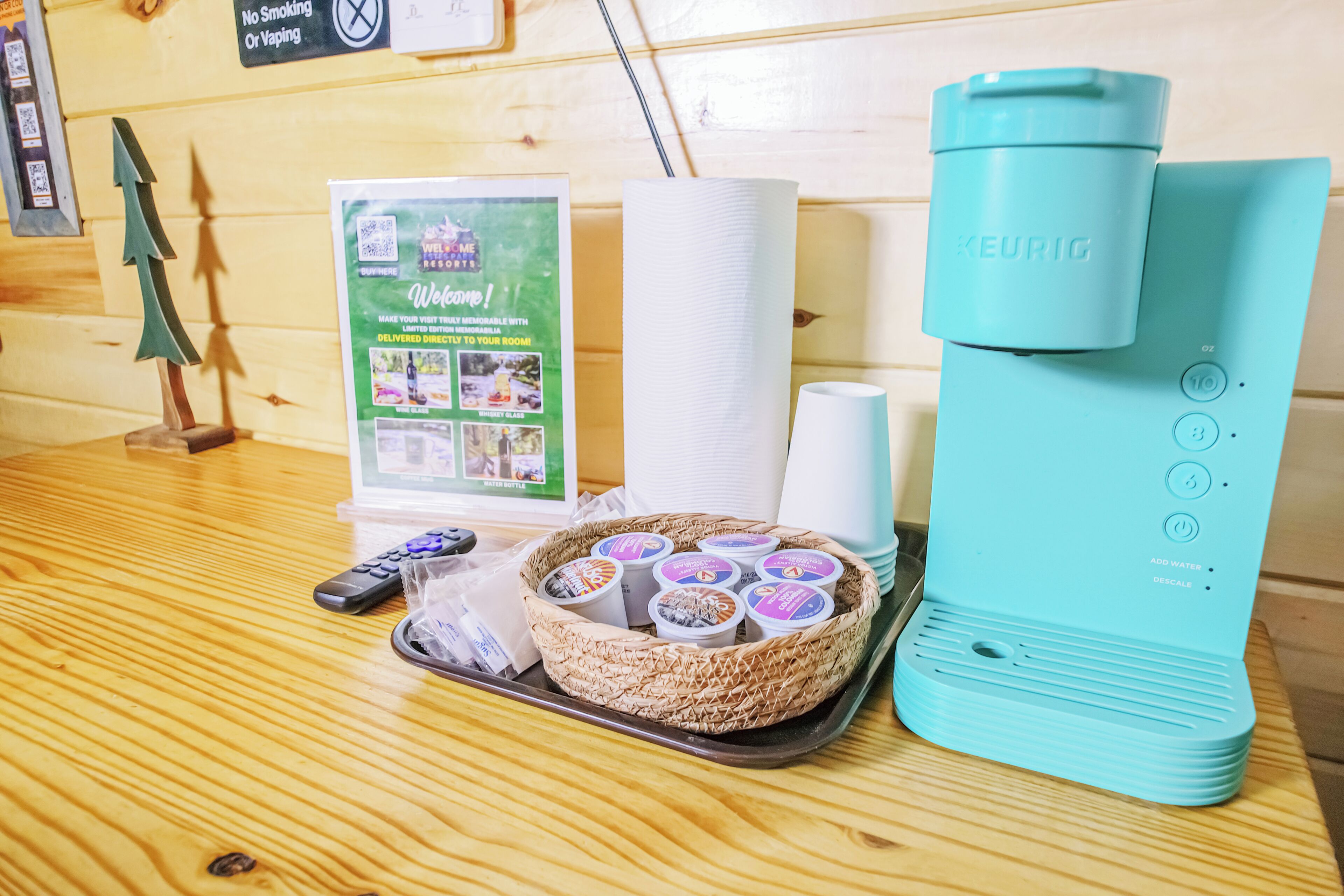 A coffee maker, a tray with coffee pods, a remote control, and a welcome sign on a wooden table.