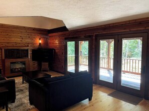Interior de uma cabana de tronco com lareira, televisão, sofá e porta de vidro deslizante que dá para um deck.